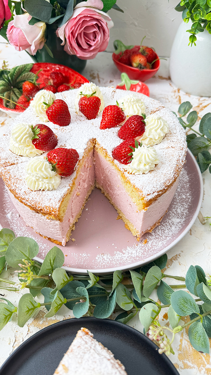 Die Käse-Sahne-Torte mit Erdbeeren hat einen luftigen, goldbraunen Biskuitboden und eine dicke, rosa Käse-Sahne-Füllung mit Erdbeergeschmack. Auf der obersten Schicht sind frische Erdbeeren und Sahnetupfer kunstvoll arrangiert, die mit einer feinen Schicht Puderzucker bestreut sind. Ein bereits geschnittenes Stück zeigt die cremige Füllung und die luftigen Schichten, die die Torte besonders einladend machen.