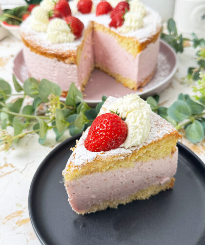 Die Käse-Sahne-Torte mit Erdbeeren hat einen luftigen, goldbraunen Biskuitboden und eine dicke, rosa Käse-Sahne-Füllung mit Erdbeergeschmack. Auf der obersten Schicht sind frische Erdbeeren und Sahnetupfer kunstvoll arrangiert, die mit einer feinen Schicht Puderzucker bestreut sind. Ein bereits geschnittenes Stück zeigt die cremige Füllung und die luftigen Schichten, die die Torte besonders einladend machen.