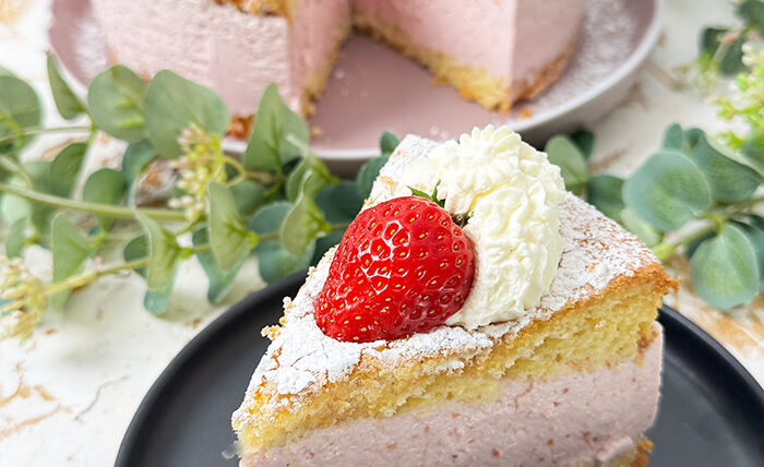 Die Käse-Sahne-Torte mit Erdbeeren hat einen luftigen, goldbraunen Biskuitboden und eine dicke, rosa Käse-Sahne-Füllung mit Erdbeergeschmack. Auf der obersten Schicht sind frische Erdbeeren und Sahnetupfer kunstvoll arrangiert, die mit einer feinen Schicht Puderzucker bestreut sind. Ein bereits geschnittenes Stück zeigt die cremige Füllung und die luftigen Schichten, die die Torte besonders einladend machen.