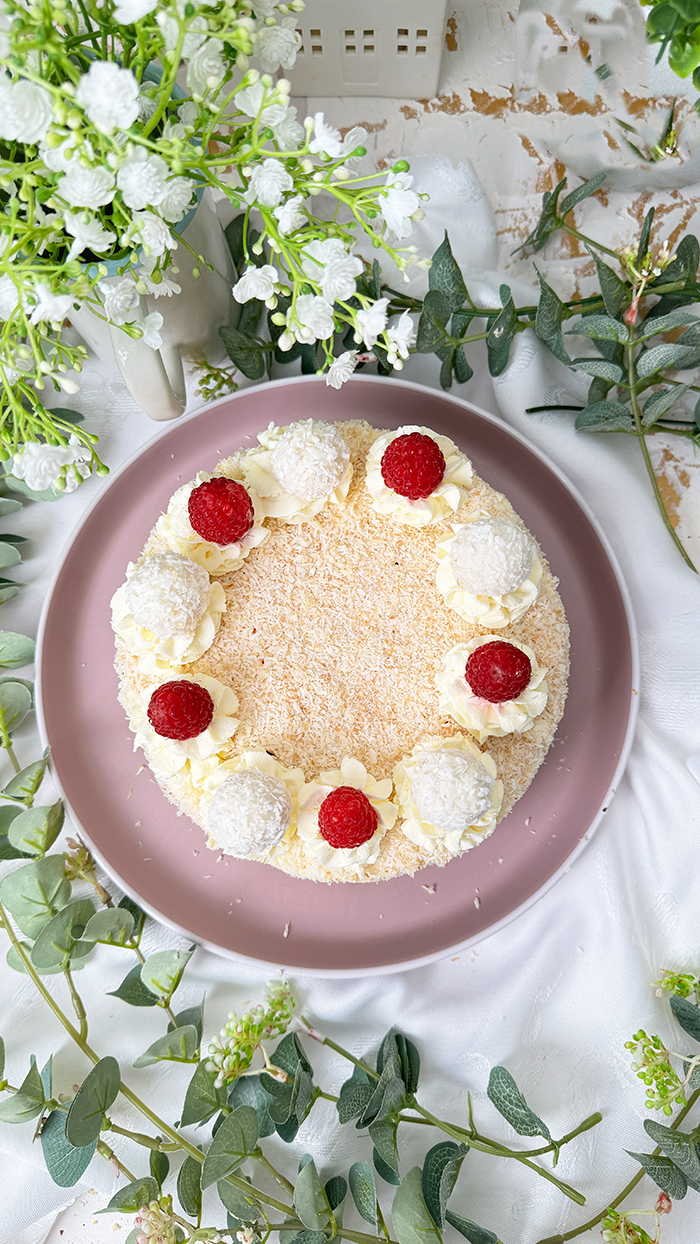 Das Bild zeigt eine Raffaello-Torte mit mehreren Schichten hellem Biskuit und cremiger Füllung, bedeckt mit Kokosraspeln. Die Torte ist dekoriert mit Raffaello-Kugeln, Himbeeren und Sahnetupfen. Ein Stück Torte liegt auf einem grauen Teller mit einer goldenen Gabel daneben, umgeben von grünen Pflanzen und weißen Blumen.