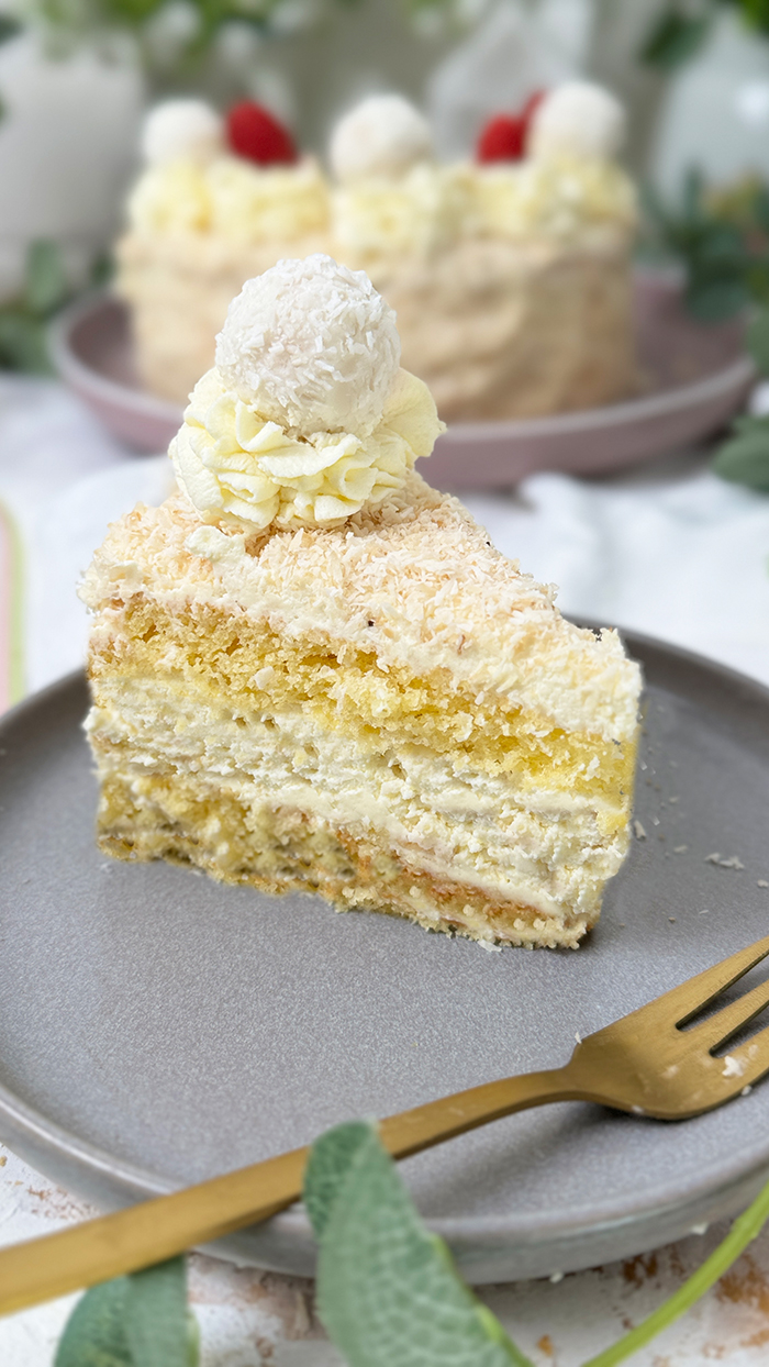 Das Bild zeigt eine Raffaello-Torte mit mehreren Schichten hellem Biskuit und cremiger Füllung, bedeckt mit Kokosraspeln. Die Torte ist dekoriert mit Raffaello-Kugeln, Himbeeren und Sahnetupfen. Ein Stück Torte liegt auf einem grauen Teller mit einer goldenen Gabel daneben, umgeben von grünen Pflanzen und weißen Blumen.