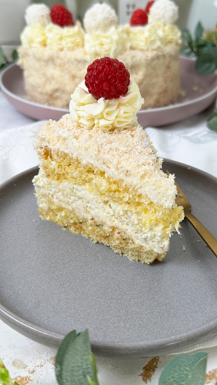 Das Bild zeigt eine Raffaello-Torte mit mehreren Schichten hellem Biskuit und cremiger Füllung, bedeckt mit Kokosraspeln. Die Torte ist dekoriert mit Raffaello-Kugeln, Himbeeren und Sahnetupfen. Ein Stück Torte liegt auf einem grauen Teller mit einer goldenen Gabel daneben, umgeben von grünen Pflanzen und weißen Blumen.