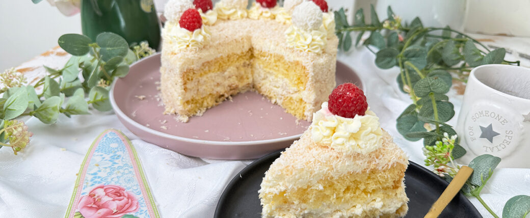 Das Bild zeigt eine Raffaello-Torte mit mehreren Schichten hellem Biskuit und cremiger Füllung, bedeckt mit Kokosraspeln. Die Torte ist dekoriert mit Raffaello-Kugeln, Himbeeren und Sahnetupfen. Ein Stück Torte liegt auf einem grauen Teller mit einer goldenen Gabel daneben, umgeben von grünen Pflanzen und weißen Blumen.