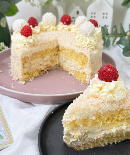 Das Bild zeigt eine Raffaello-Torte mit mehreren Schichten hellem Biskuit und cremiger Füllung, bedeckt mit Kokosraspeln. Die Torte ist dekoriert mit Raffaello-Kugeln, Himbeeren und Sahnetupfen. Ein Stück Torte liegt auf einem grauen Teller mit einer goldenen Gabel daneben, umgeben von grünen Pflanzen und weißen Blumen.