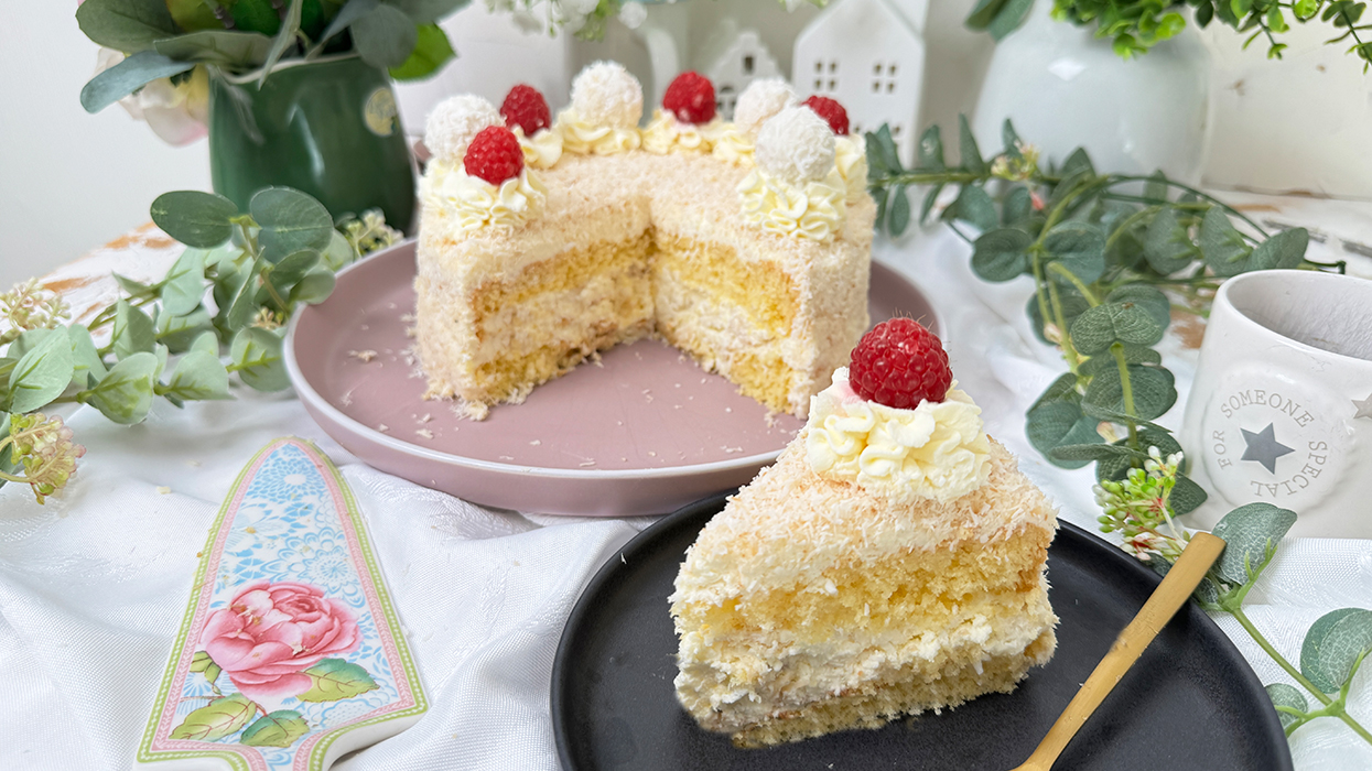 Das Bild zeigt eine Raffaello-Torte mit mehreren Schichten hellem Biskuit und cremiger Füllung, bedeckt mit Kokosraspeln. Die Torte ist dekoriert mit Raffaello-Kugeln, Himbeeren und Sahnetupfen. Ein Stück Torte liegt auf einem grauen Teller mit einer goldenen Gabel daneben, umgeben von grünen Pflanzen und weißen Blumen.