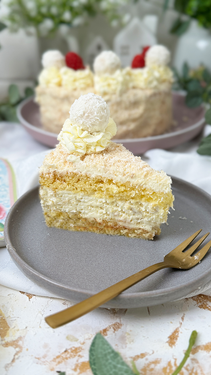 Das Bild zeigt eine Raffaello-Torte mit mehreren Schichten hellem Biskuit und cremiger Füllung, bedeckt mit Kokosraspeln. Die Torte ist dekoriert mit Raffaello-Kugeln, Himbeeren und Sahnetupfen. Ein Stück Torte liegt auf einem grauen Teller mit einer goldenen Gabel daneben, umgeben von grünen Pflanzen und weißen Blumen.