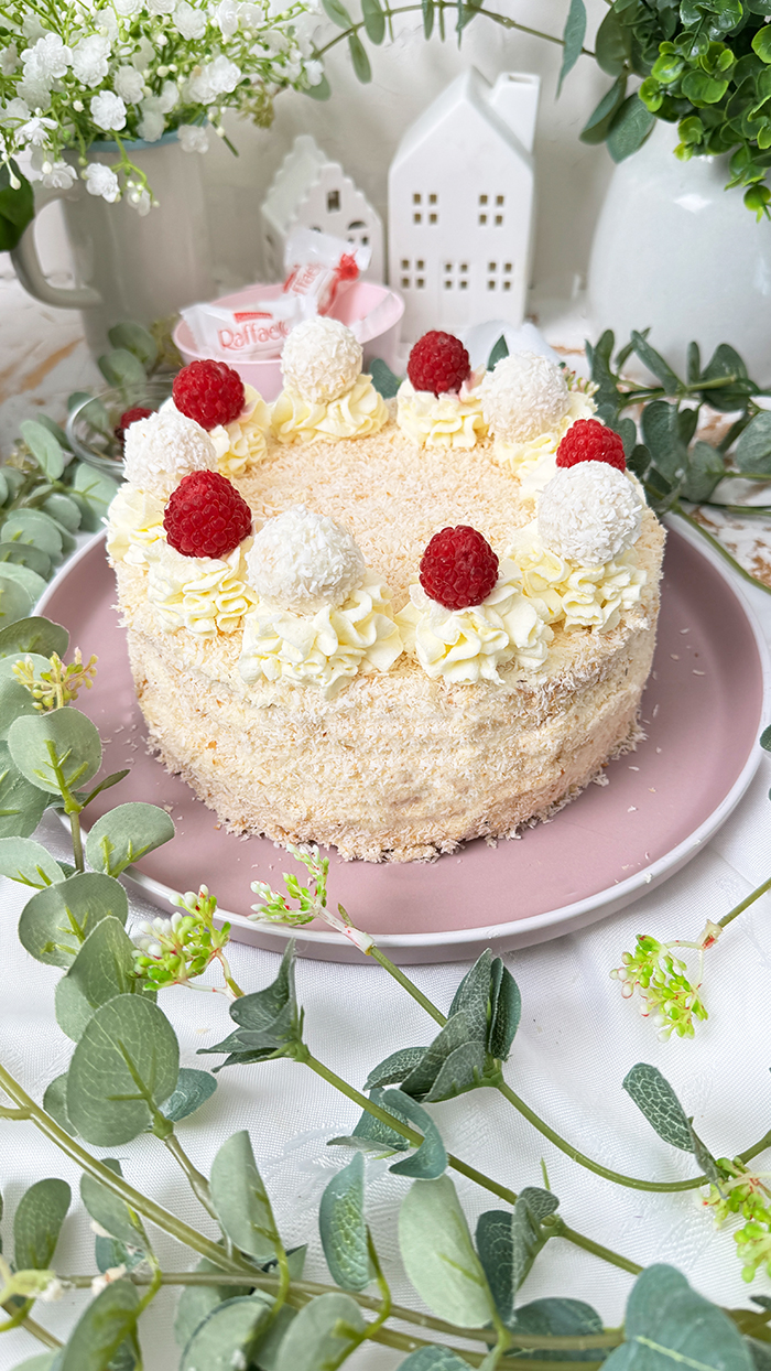 Das Bild zeigt eine Raffaello-Torte mit mehreren Schichten hellem Biskuit und cremiger Füllung, bedeckt mit Kokosraspeln. Die Torte ist dekoriert mit Raffaello-Kugeln, Himbeeren und Sahnetupfen. Ein Stück Torte liegt auf einem grauen Teller mit einer goldenen Gabel daneben, umgeben von grünen Pflanzen und weißen Blumen.