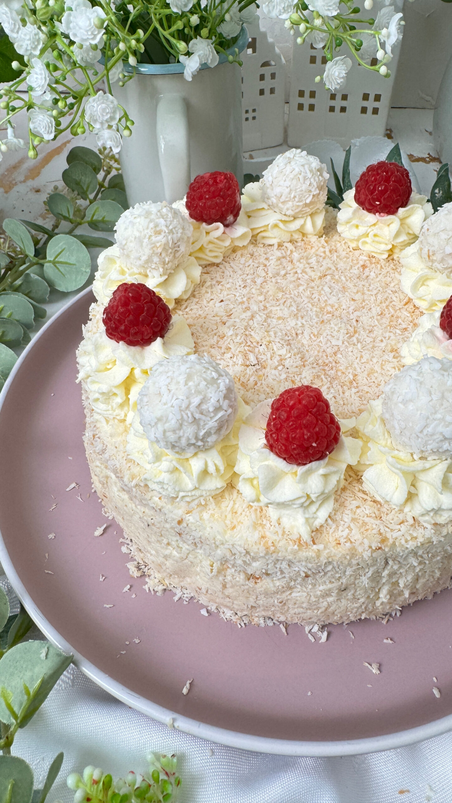 Das Bild zeigt eine Raffaello-Torte mit mehreren Schichten hellem Biskuit und cremiger Füllung, bedeckt mit Kokosraspeln. Die Torte ist dekoriert mit Raffaello-Kugeln, Himbeeren und Sahnetupfen. Ein Stück Torte liegt auf einem grauen Teller mit einer goldenen Gabel daneben, umgeben von grünen Pflanzen und weißen Blumen.