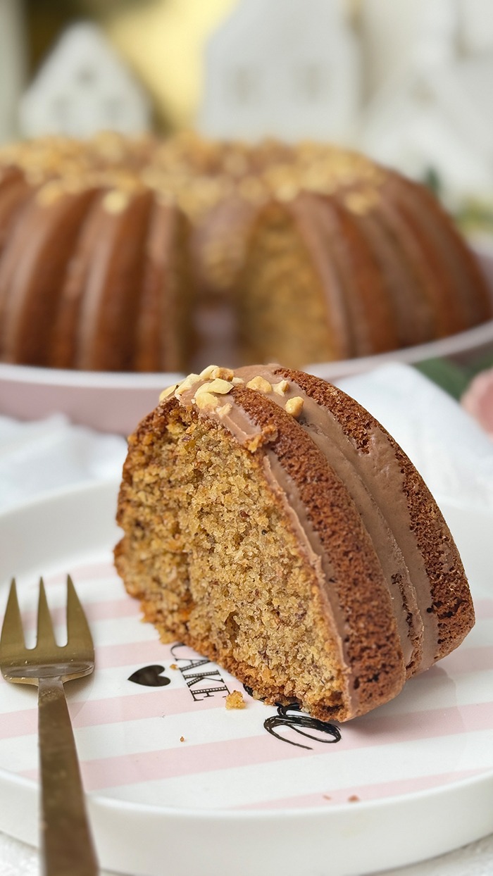 Ein saftiger nusskuchen im gugelhupf-stil mit haselnussglasur und gehackten nüssen liegt auf einem dekorativen teller, während der gesamte kuchen im hintergrund präsentiert wird.