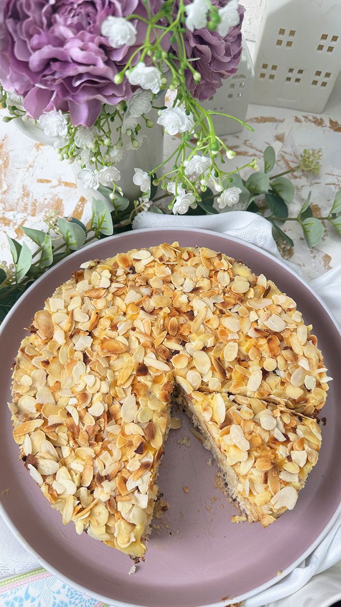 Auf dem Bild sieht man ein Stück Schwedische Mandeltorte, das auf einem grauen Teller angerichtet ist. Die Torte besteht aus mehreren Schichten: ein luftiger Mandelboden, dazwischen eine reichhaltige Vanillebuttercreme und obenauf eine Schicht gerösteter Mandelblättchen.