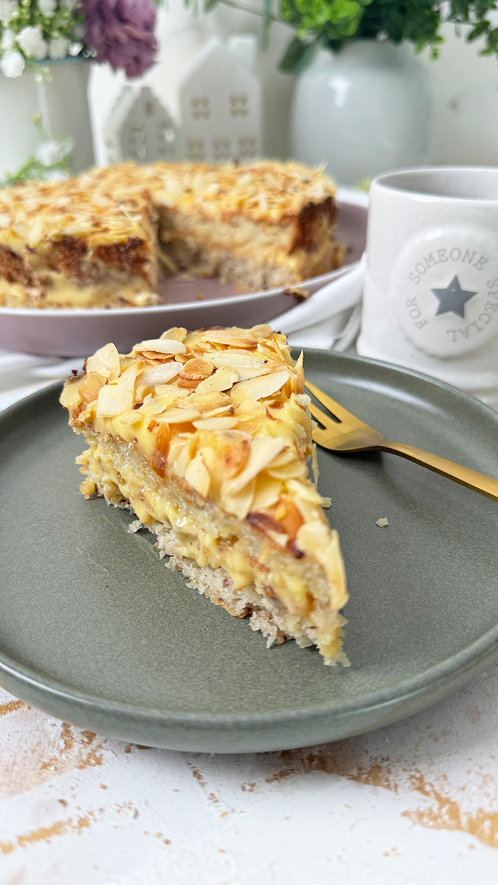 Auf dem Bild sieht man ein Stück Schwedische Mandeltorte, das auf einem grauen Teller angerichtet ist. Die Torte besteht aus mehreren Schichten: ein luftiger Mandelboden, dazwischen eine reichhaltige Vanillebuttercreme und obenauf eine Schicht gerösteter Mandelblättchen.