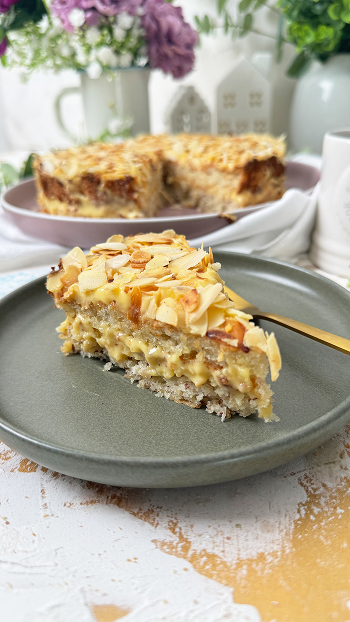 Auf dem Bild sieht man ein Stück Schwedische Mandeltorte, das auf einem grauen Teller angerichtet ist. Die Torte besteht aus mehreren Schichten: ein luftiger Mandelboden, dazwischen eine reichhaltige Vanillebuttercreme und obenauf eine Schicht gerösteter Mandelblättchen.