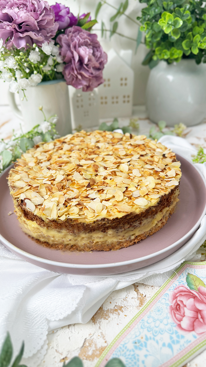 Auf dem Bild sieht man ein Stück Schwedische Mandeltorte, das auf einem grauen Teller angerichtet ist. Die Torte besteht aus mehreren Schichten: ein luftiger Mandelboden, dazwischen eine reichhaltige Vanillebuttercreme und obenauf eine Schicht gerösteter Mandelblättchen.