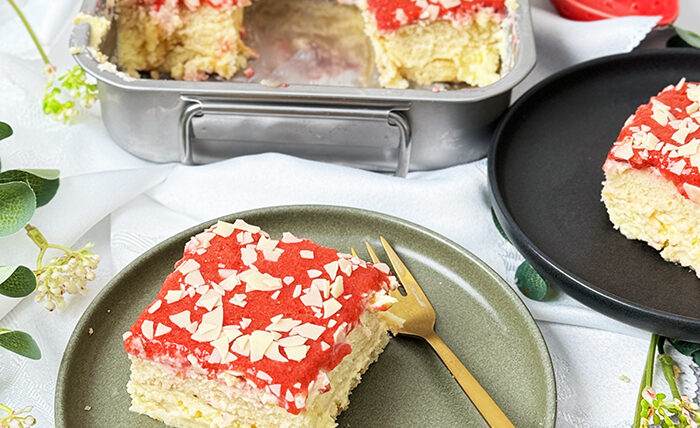 Dieses Bild zeigt ein Spaghettieis-Tiramisu. Das Dessert befindet sich in einer Backform, aus der ein quadratisches Stück herausgeschnitten und auf einem Teller angerichtet wurde, bereit zum Verzehr. Die oberste Schicht ist mit einer Erdbeerglasur und weißen Schokoladenraspeln garniert. Das Setting ist hell und mit Blumen und kleinen Pflanzen dekoriert, was auf ein Frühlings- oder Sommerthema hindeutet.