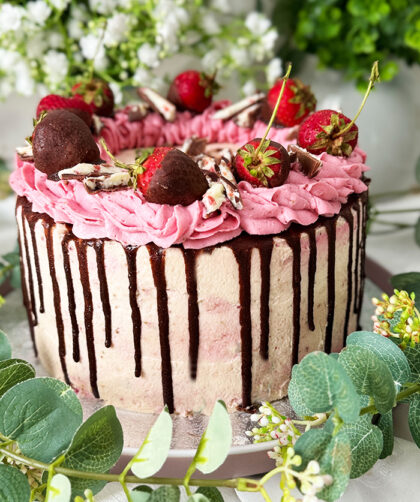 Das Bild zeigt eine zweischichtige Yogurette-Torte, dekoriert mit rosa Sahne, Schokoladen-Drip, in Schokolade getauchten Erdbeeren und Yogurette-Stückchen, umgeben von Blumen. Sommertorte