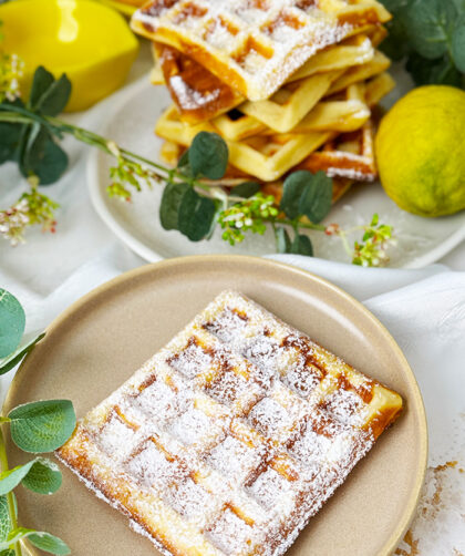 Ein Teller mit einer Zitronen-Quark-Waffel, bestreut mit Puderzucker, steht im Vordergrund, während im Hintergrund ein Stapel weiterer Waffeln neben Zitronen und einer Tasse zu sehen ist.