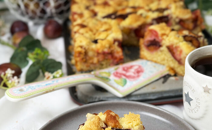 Auf dem Bilde erblicket man einen köstlichen Zwetschgen-Streuselkuchen mit goldbrauner Streuseldecke und saftigen Früchten. Ein Stück liegt auf einem Teller, während der restliche Kuchen im Hintergrund in der Form ruht, umrahmt von zarter grüner Dekoration. Ein wahrhaft verlockender Anblick!